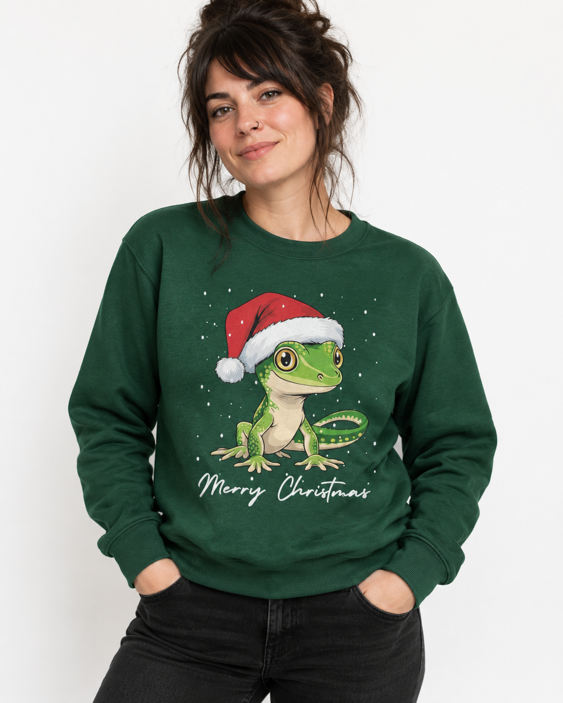 A woman wearing a dark green crewneck sweatshirt with a graphic of a green gecko wearing a red Santa hat and the text 'Merry Christmas' underneath, paired with black jeans against a plain white background.