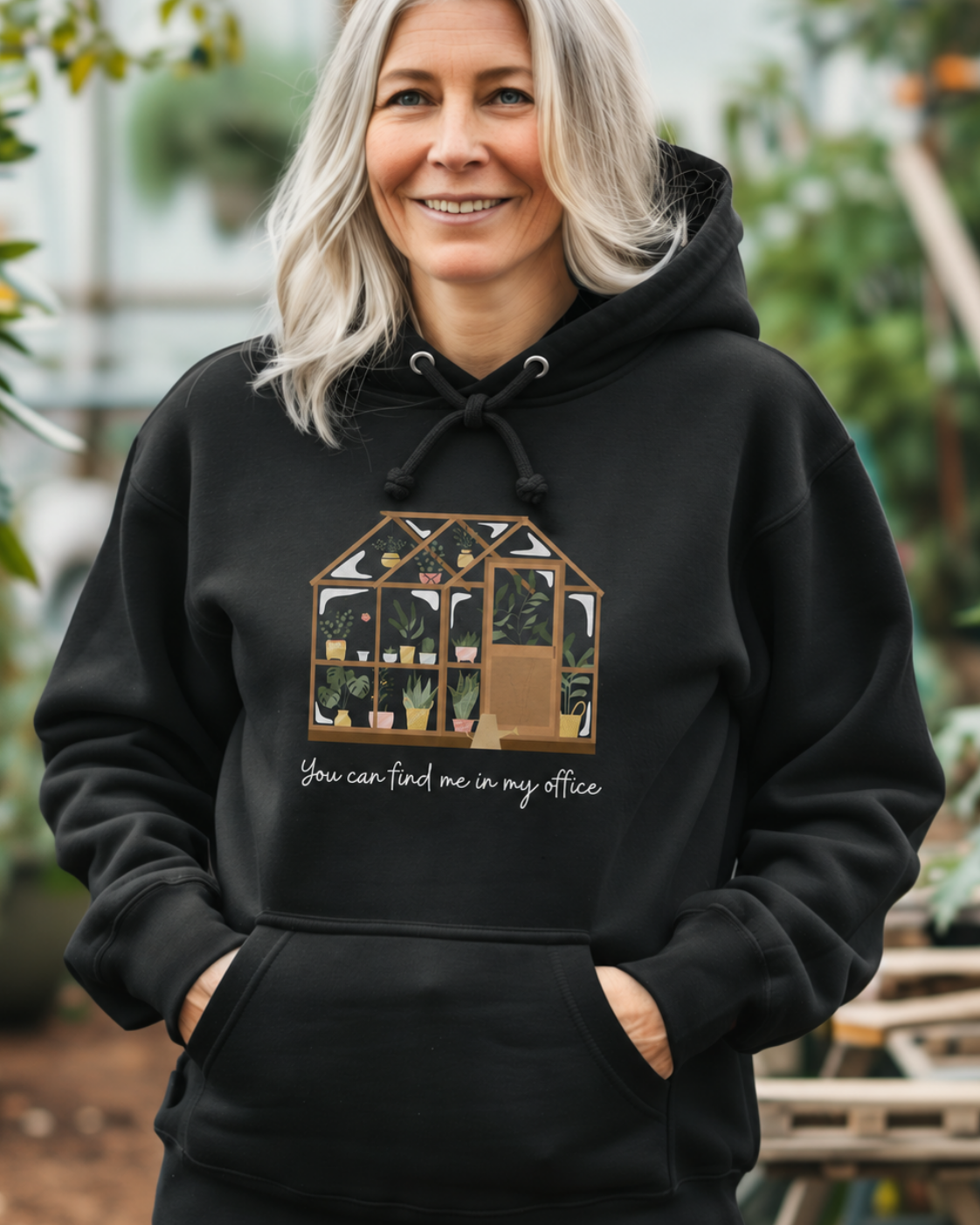 Woman wearing a black greenhouse hoodie with “You can find me in my office” print while standing among plants.