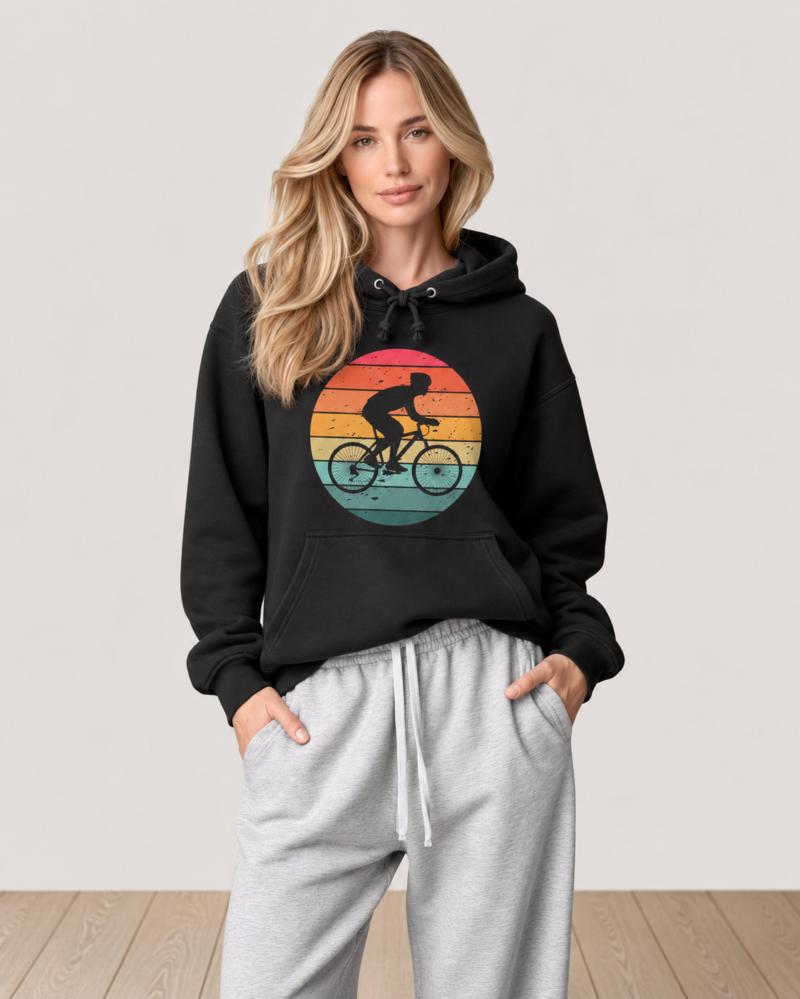 A woman modeling a black pullover hoodie with a retro circular bicycle graphic in sunset colors, paired with light gray sweatpants, against a neutral studio background.