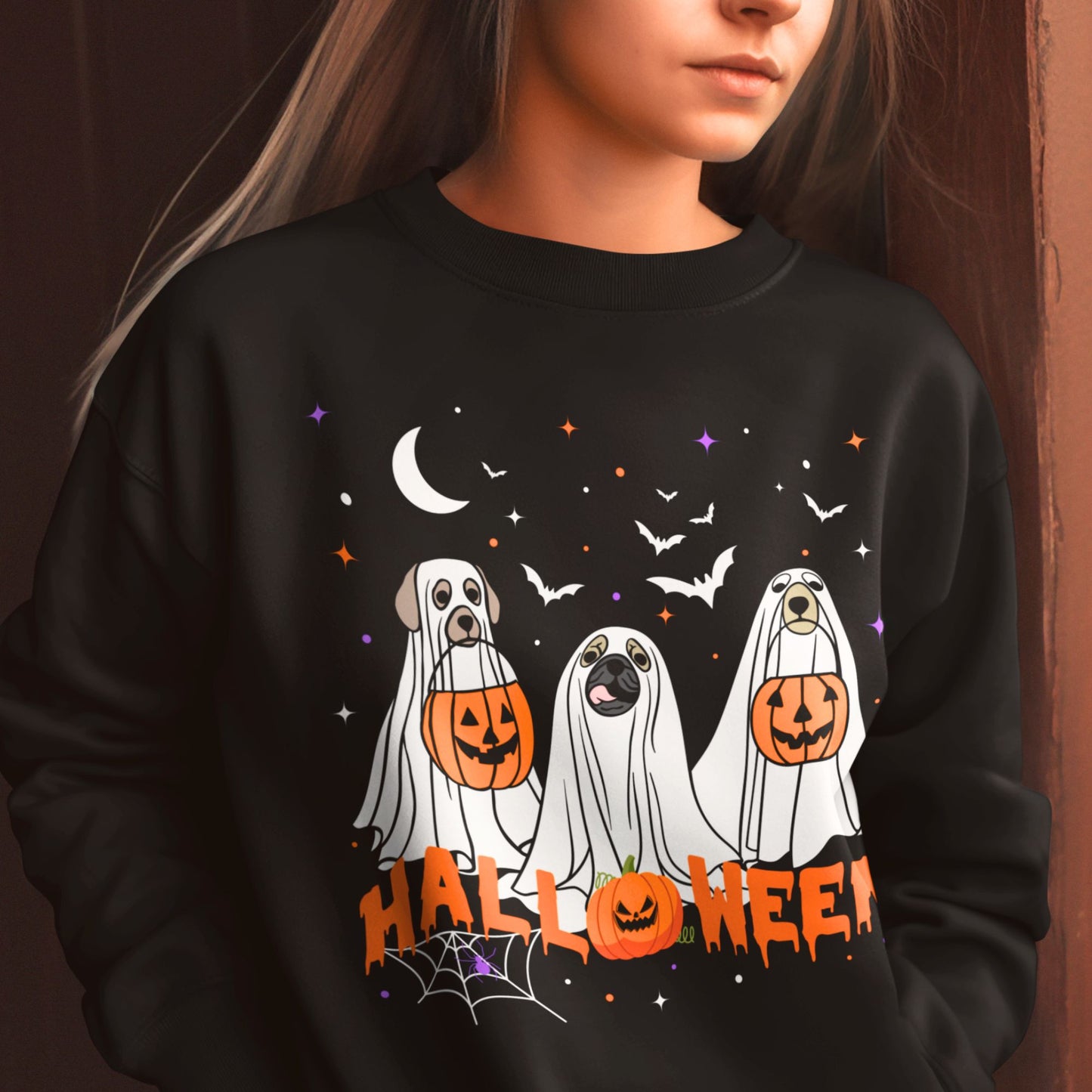 Woman wears black Halloween sweatshirt featuring three dogs in ghost costumes holding pumpkin heads, with bats, stars, and "HALLOWEEN" text.