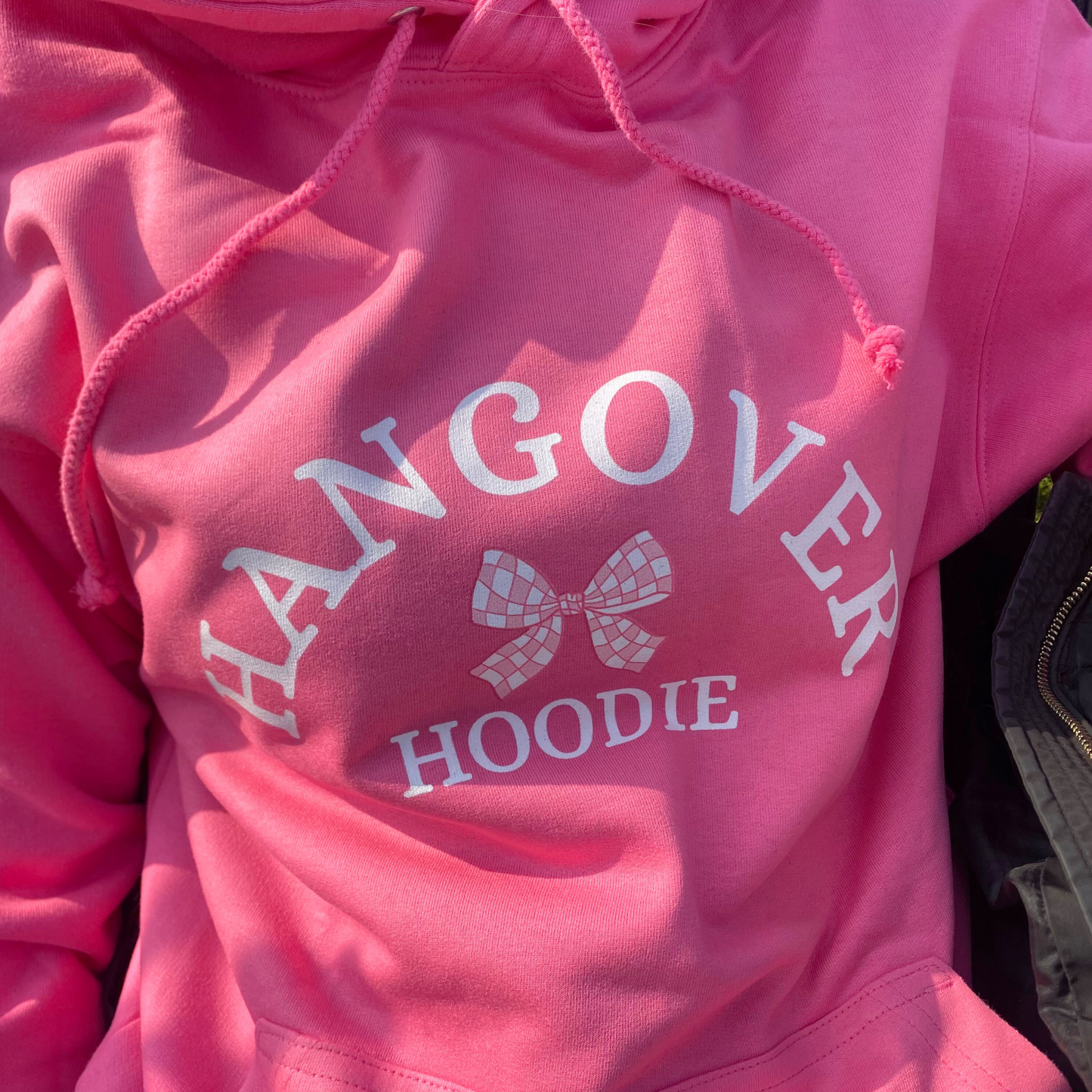 A pink hoodie with the text 'HANGOVER HOODIE' printed on the front, accompanied by a graphic of a checkered bow.