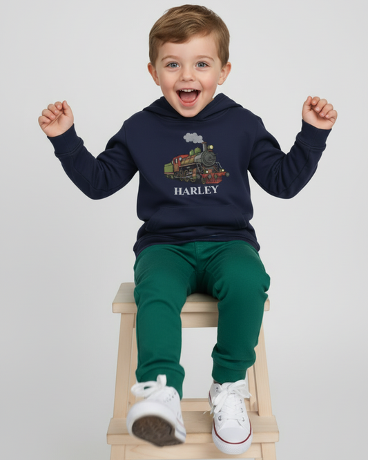 Smiling child model sitting on a stool wearing a navy personalised steam train hoodie, green joggers and white trainers in a bright studio setting.