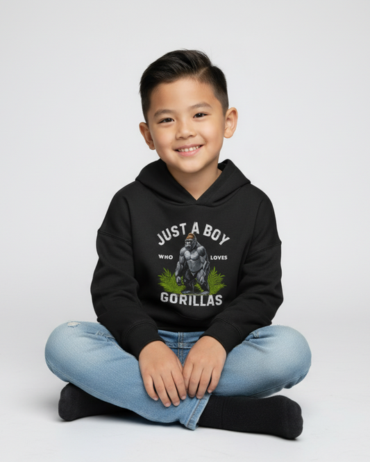 Smiling young boy sitting cross-legged wearing a black “Just A Boy Who Loves Gorillas” hoodie on a light studio background