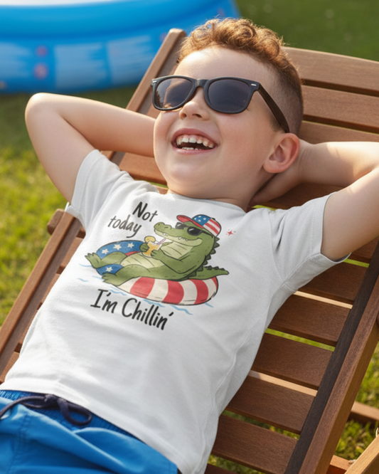 A young boy wearing black sunglasses and a white graphic t-shirt with a green alligator design, relaxing on a wooden deck chair in a backyard with a blue pool visible in the background.