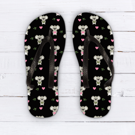Pair of kids black flip flops featuring a playful koala pattern with pink hearts and green leaves, displayed on a white wooden background.