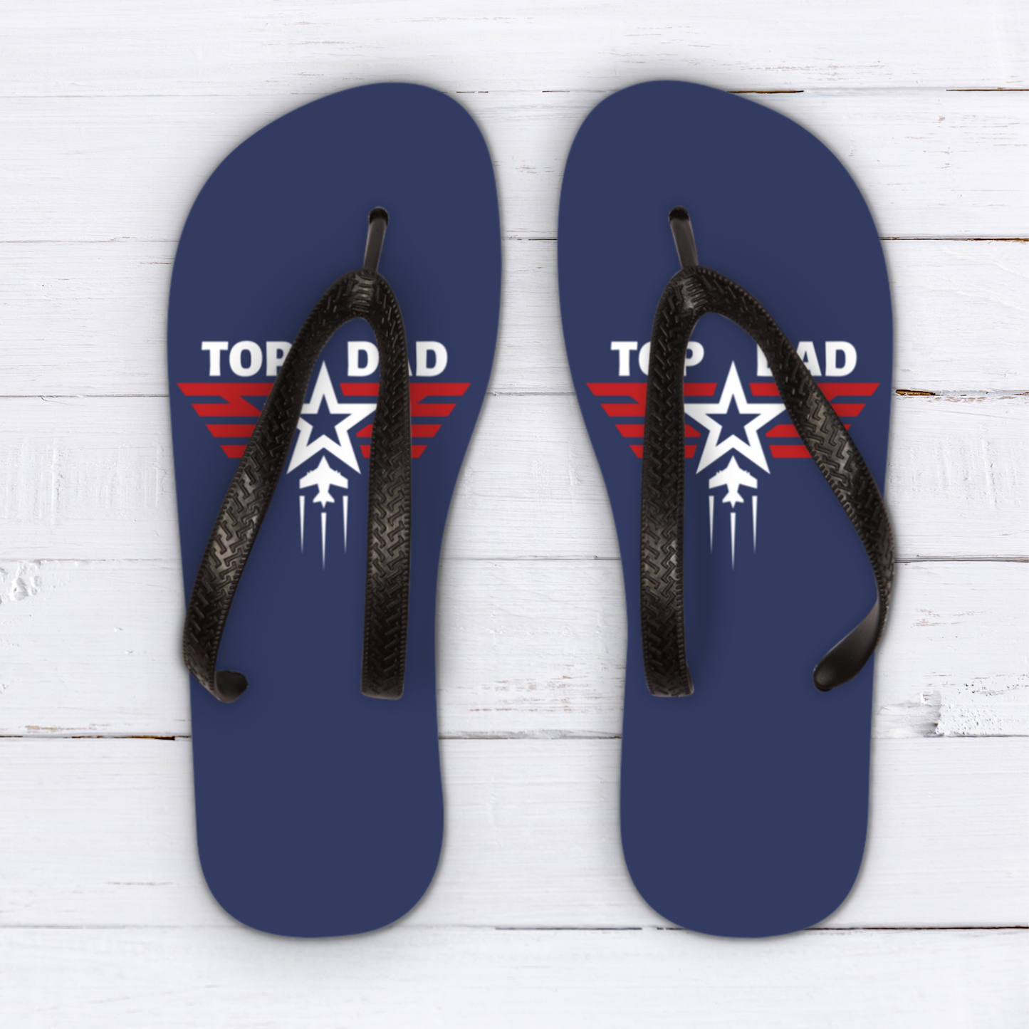Top Dad Flip Flops: Two blue flip-flops with black straps are displayed on a white wooden surface. Each flip-flop features a red and white graphic resembling a fighter jet with the text "TOP DAD" on the left flip-flop and "TOP DAD" on the right.