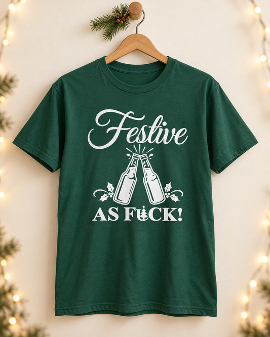 A dark green short-sleeved adult t-shirt hanging on a wooden hanger against a neutral wall with a pine sprig and string lights, featuring a white graphic of two clinking beer bottles, holly leaf accents, and the text "FESTIVE AS FUCK!" with a tiny Christmas tree.