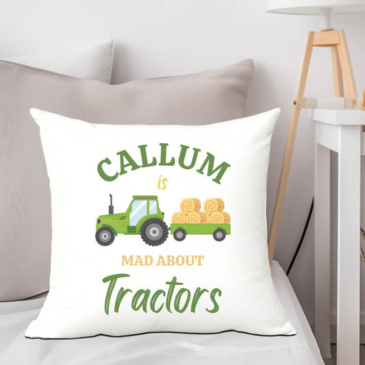 A white canvas cushion propped up on a bed, the cushion cover has a printed design of a green tractor and trailer with bales of hay and the wording "Callum is mad about Tractors"