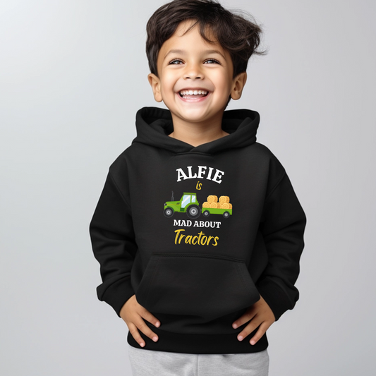 A little boy wearing a black hoodie with a printed tractor and words saying "Alfie is mad about tractors"