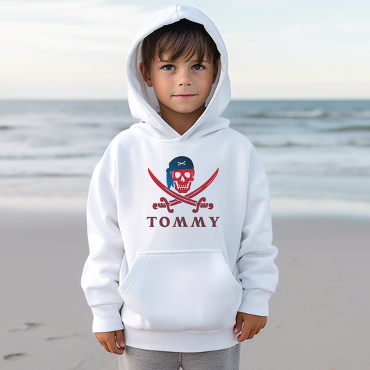 A little boy wearing a white hoodie with a printed pirate skull, swords and a  personalised name