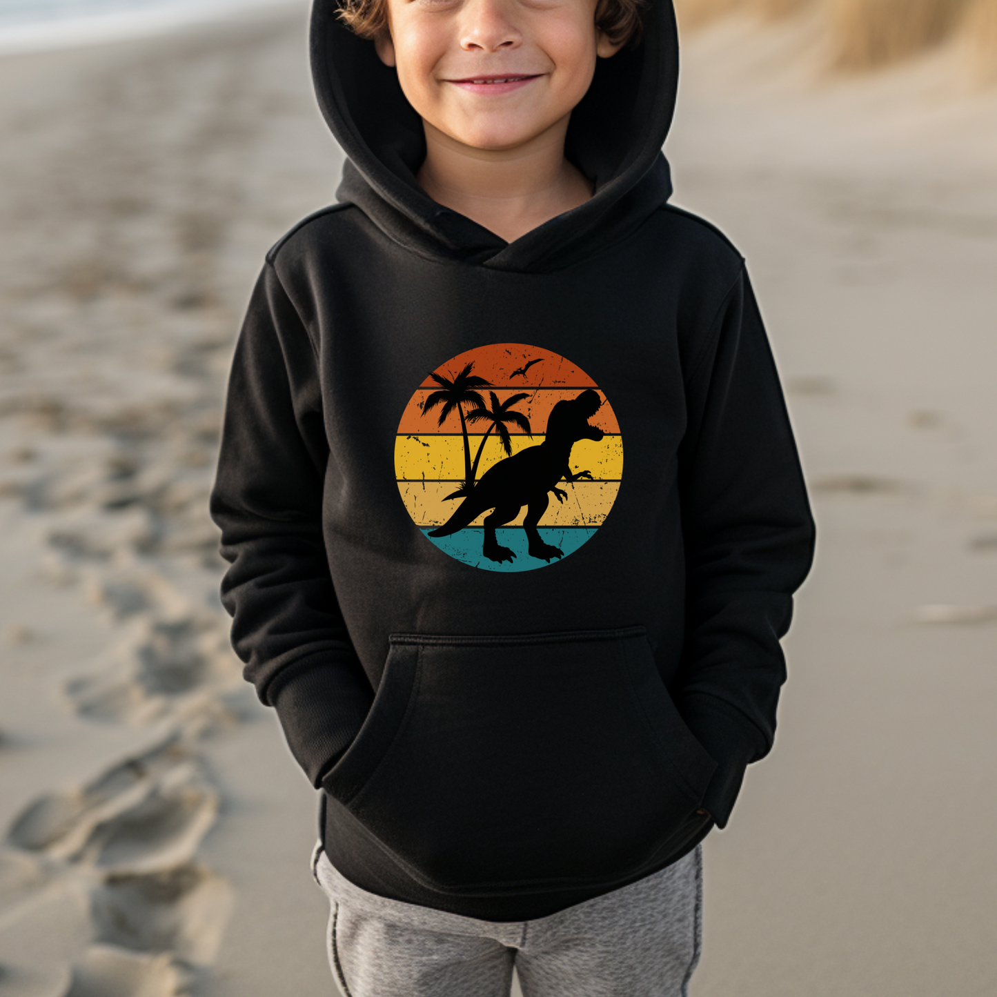 Boy standing on a beach wearing a black hoodie with a retro sunset t-rex silhouette and palm tree printed design.