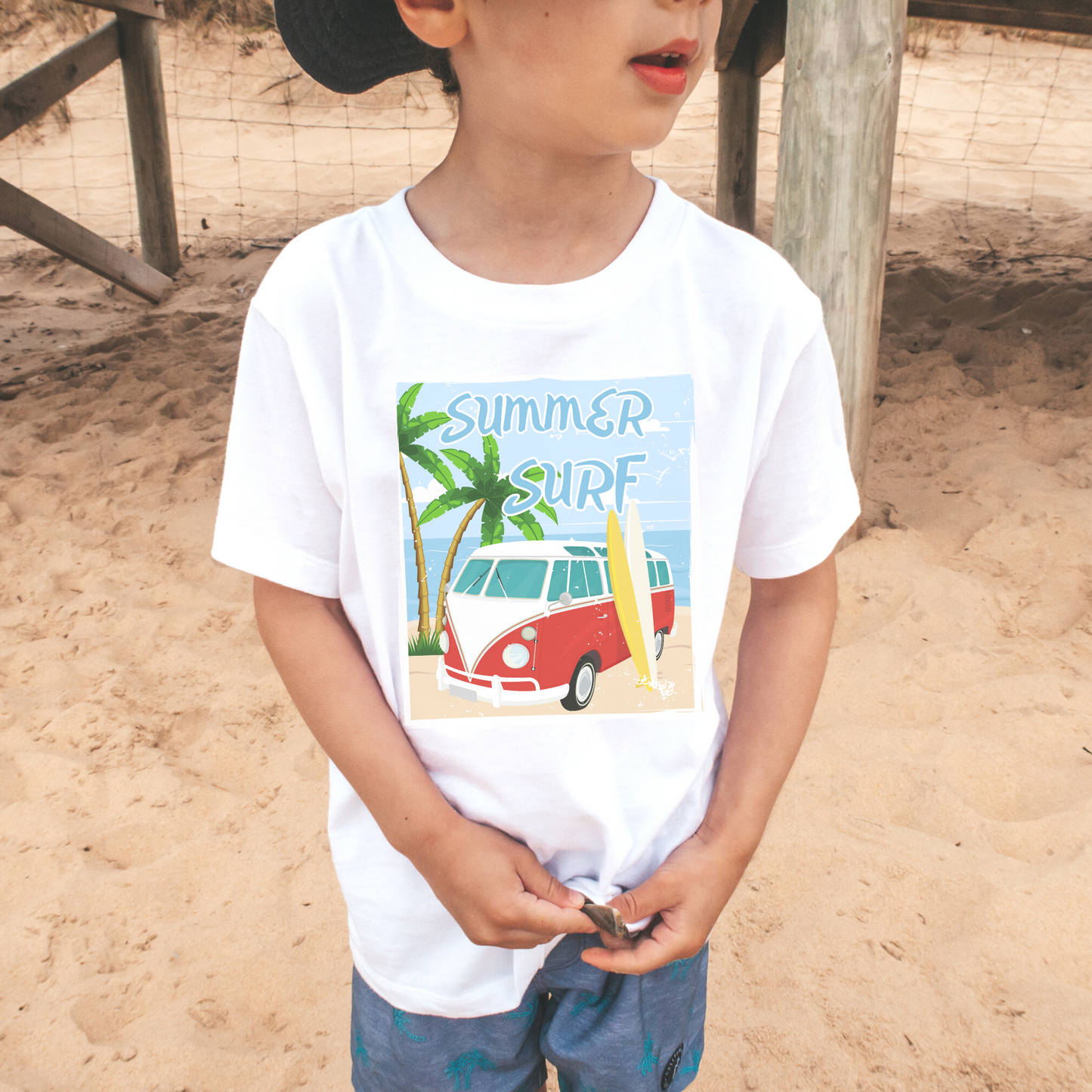 A little boy wearing a white short sleeve t-shirt with a printed camper van and surf board graphic with the words "summer surf"