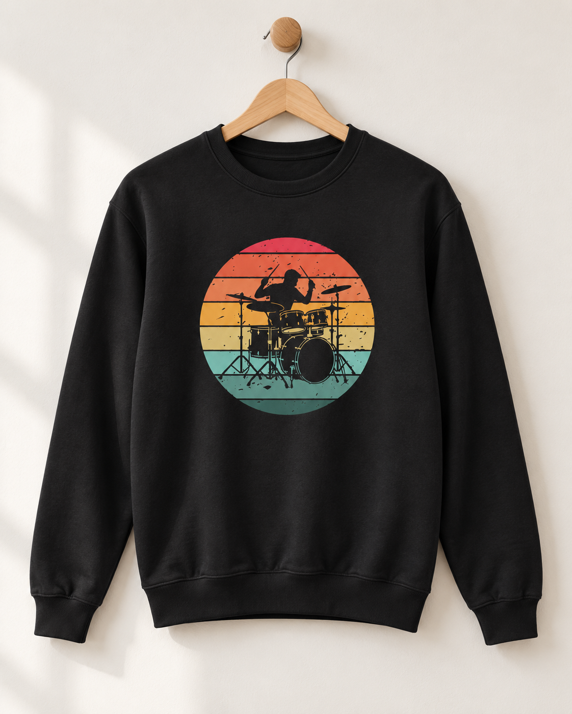 Black adult sweatshirt hanging from a wooden wall hook, featuring a drummer and drum kit silhouette against colourful distressed sunset stripes.