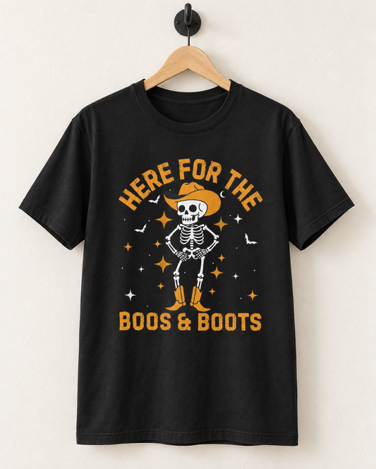Black “Here for the Boos & Boots” cowboy skeleton T-shirt displayed on a wooden hanger and black wall hook.