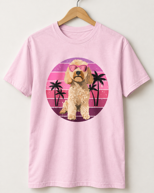 A light pink crewneck t-shirt featuring a retro-style circular graphic of a fluffy cream-coloured cockapoo dog wearing pink sunglasses, set against a pink and purple striped sunset with black palm tree silhouettes.