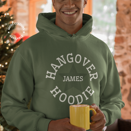 A young man standing in front of a Christmas tree holding a mug while wearing a earthy green hoodie with printed design of "Hangover Hoodie" with the name James on it