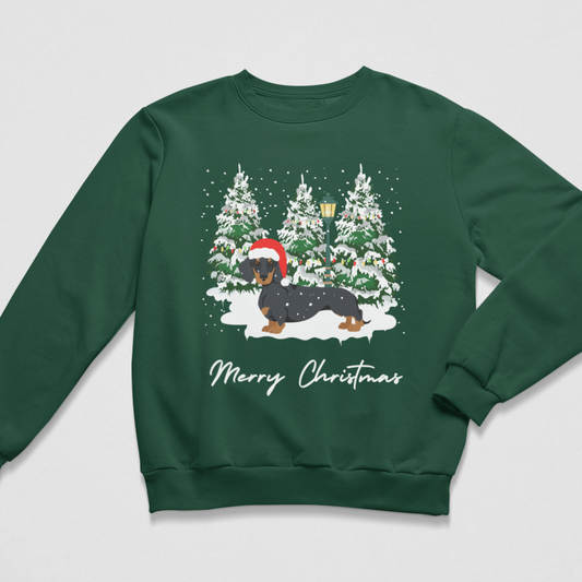A dark green crewneck sweatshirt featuring a festive Christmas graphic of a dachshund wearing a Santa hat, standing in front of snow-covered pine trees, with 'Merry Christmas' written below in white script.