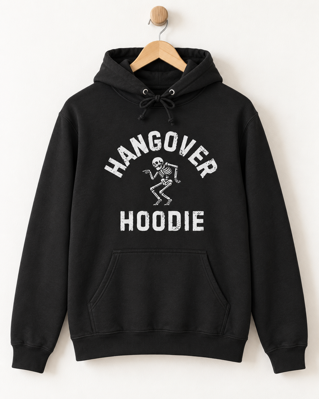 A black pullover hoodie hanging on a wooden hanger against a plain white wall. The hoodie features a white graphic print on the front with the words 'HANGOVER HOODIE' and a small dancing skeleton.