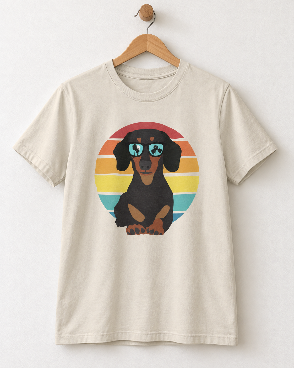 A cream-colored short-sleeve t-shirt hanging on a wooden hanger against a white wall. The shirt features a graphic print of a black and tan dachshund wearing blue sunglasses with shamrock lenses, set against a retro striped sunset background.