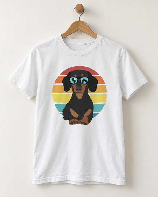 A white short-sleeve t-shirt hanging on a wooden hanger against a light beige wall. The shirt features a graphic print of a black and tan dachshund wearing blue star-shaped sunglasses, set against a retro circular sunset background with red, orange, yellow, green, and blue stripes.