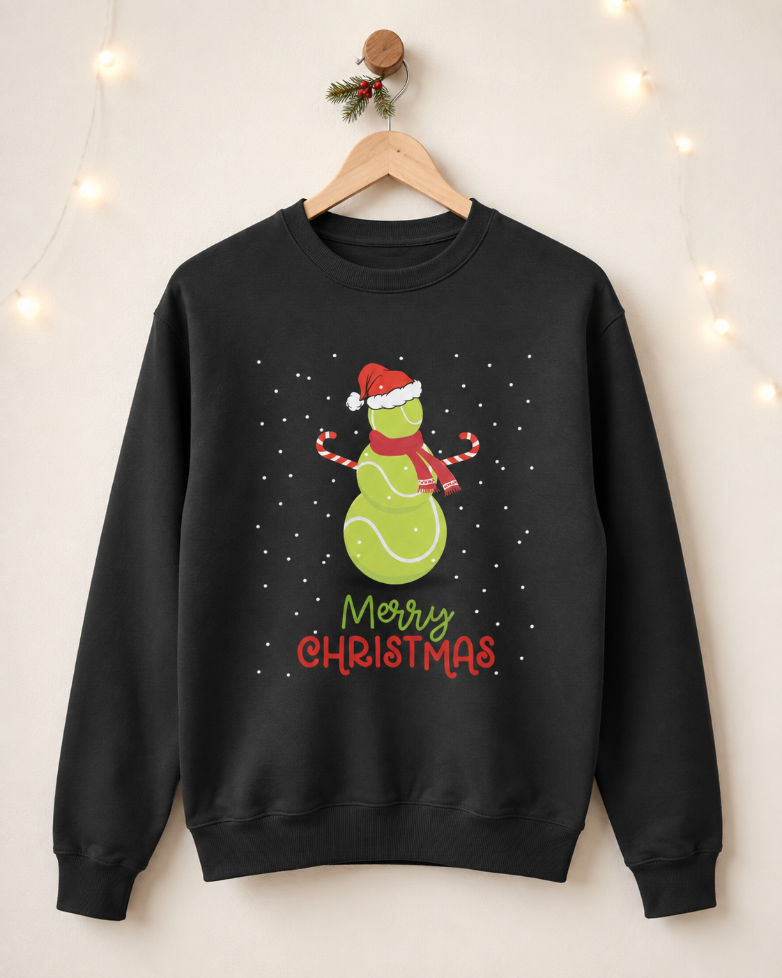 Adults black crewneck sweatshirt hanging on a wooden hanger against a warm beige wall with soft string lights. The sweatshirt features a festive graphic tennis ball snowman with a Santa hat and scarf, surrounded by white snowflakes, with 'Merry Christmas' written below in green and red lettering.