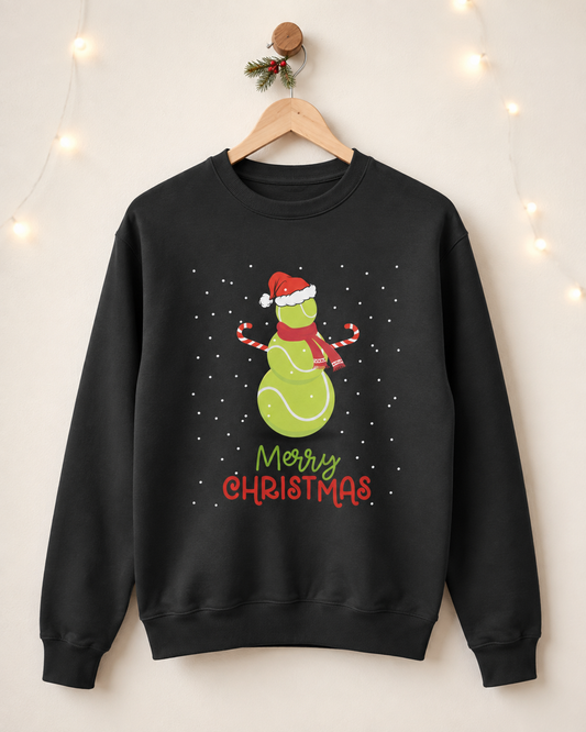 Adults black crewneck sweatshirt hanging on a wooden hanger against a warm beige wall with soft string lights. The sweatshirt features a festive graphic tennis ball snowman with a Santa hat and scarf, surrounded by white snowflakes, with 'Merry Christmas' written below in green and red lettering.