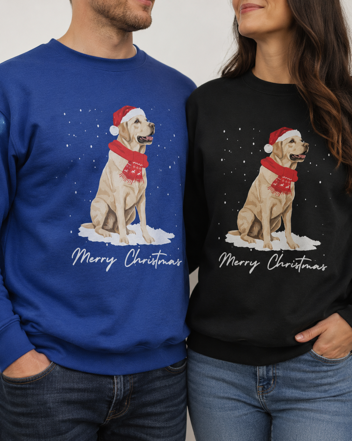 Two people wearing matching Christmas sweatshirts, one royal blue and one black, both featuring a graphic of a yellow Labrador dog wearing a Santa hat and scarf with 'Merry Christmas' written below.