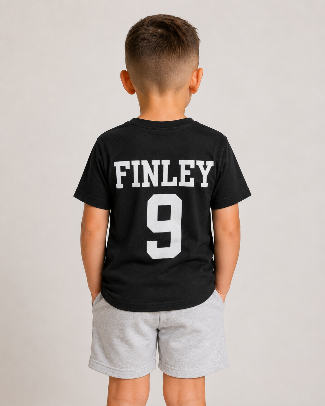 A young boy viewed from behind wearing a black t-shirt with 'FINLEY' and the number '9' printed in white on the back, paired with light gray shorts, against a plain light gray background.
