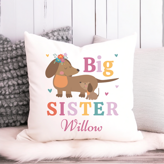 A white cushion cover featuring illustrations of two dachshunds and the text 'Big Sister' with a personalized name 'Willow'. The cover is placed on a sofa with a grey textured throw.
