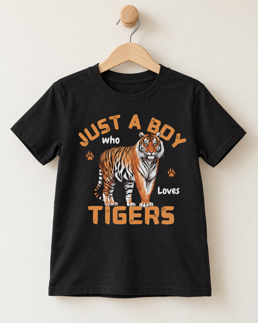 A black short-sleeve kids' t-shirt hanging on a wooden hanger against a light beige wall. The shirt features a graphic print of a tiger and the text 'JUST A BOY who Loves TIGERS' in orange and white.