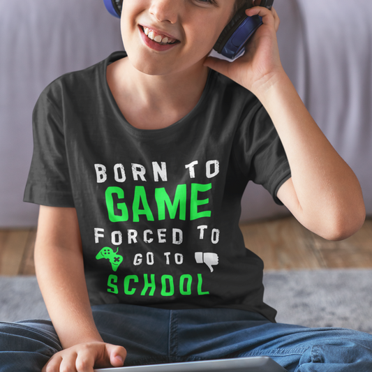 A smiling child wearing blue headphones models a black t-shirt with the text "BORN TO GAME" in white and bright green, followed by "FORCED TO GO TO SCHOOL" with a green game controller and a white thumbs-down graphic.