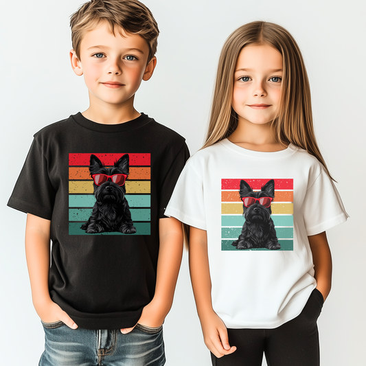 Two young children, a boy and a girl, stand side-by-side modelling t-shirts. The boy wears a black tee, and the girl wears a white one, both featuring a graphic of a black Scottish Terrier wearing red sunglasses against a retro striped background.