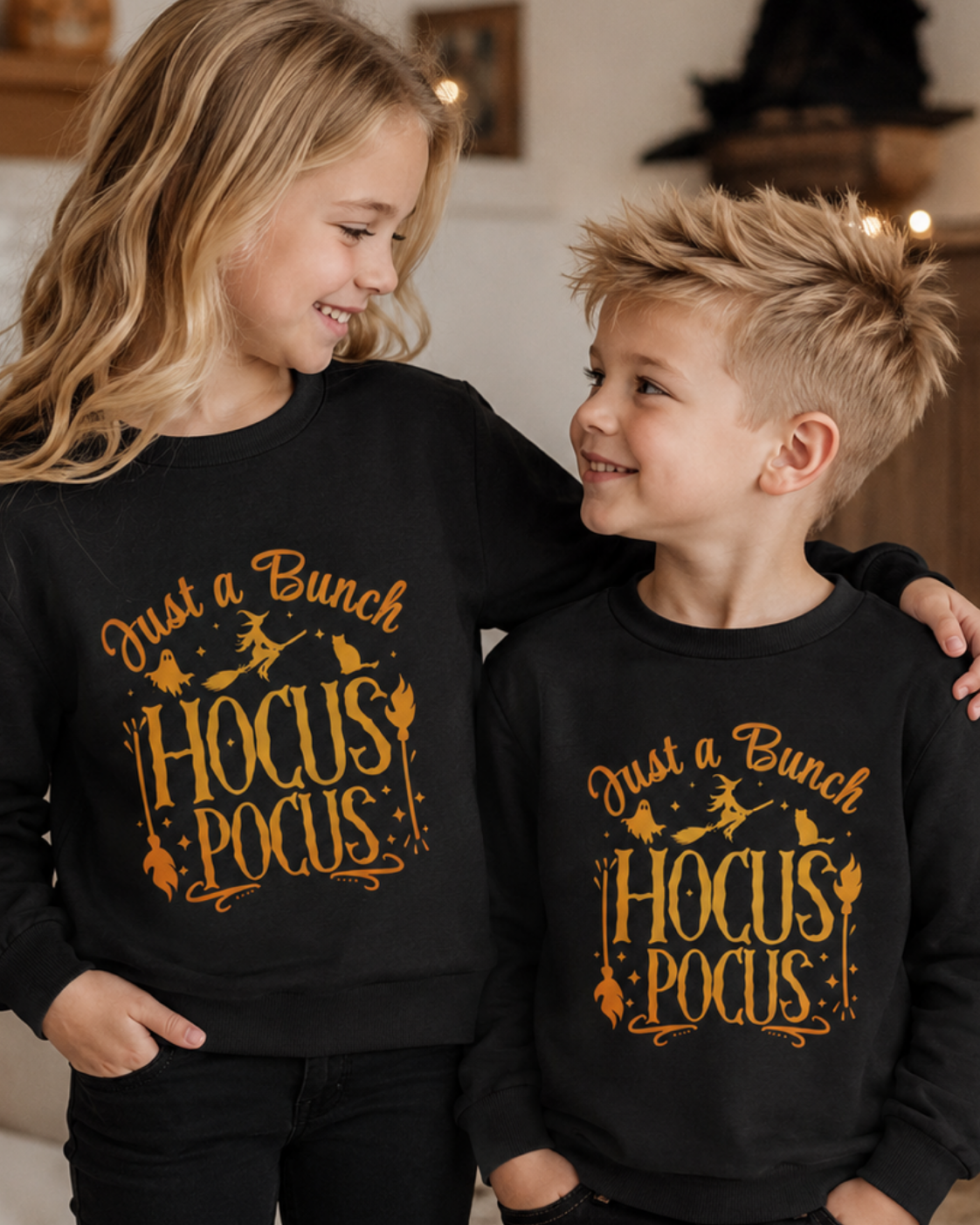 A girl and a boy wearing matching black long-sleeve sweatshirts with a golden orange graphic that reads 'Just a Bunch Hocus Pocus' with witch hats, broomsticks, and cauldron illustrations, posed indoors.
