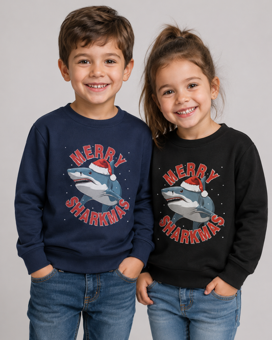 Two children standing side by side wearing matching Christmas-themed sweatshirts with a shark graphic and the text 'MERRY SHARKMAS' in red, one navy blue and one black, paired with blue jeans against a light grey studio background.