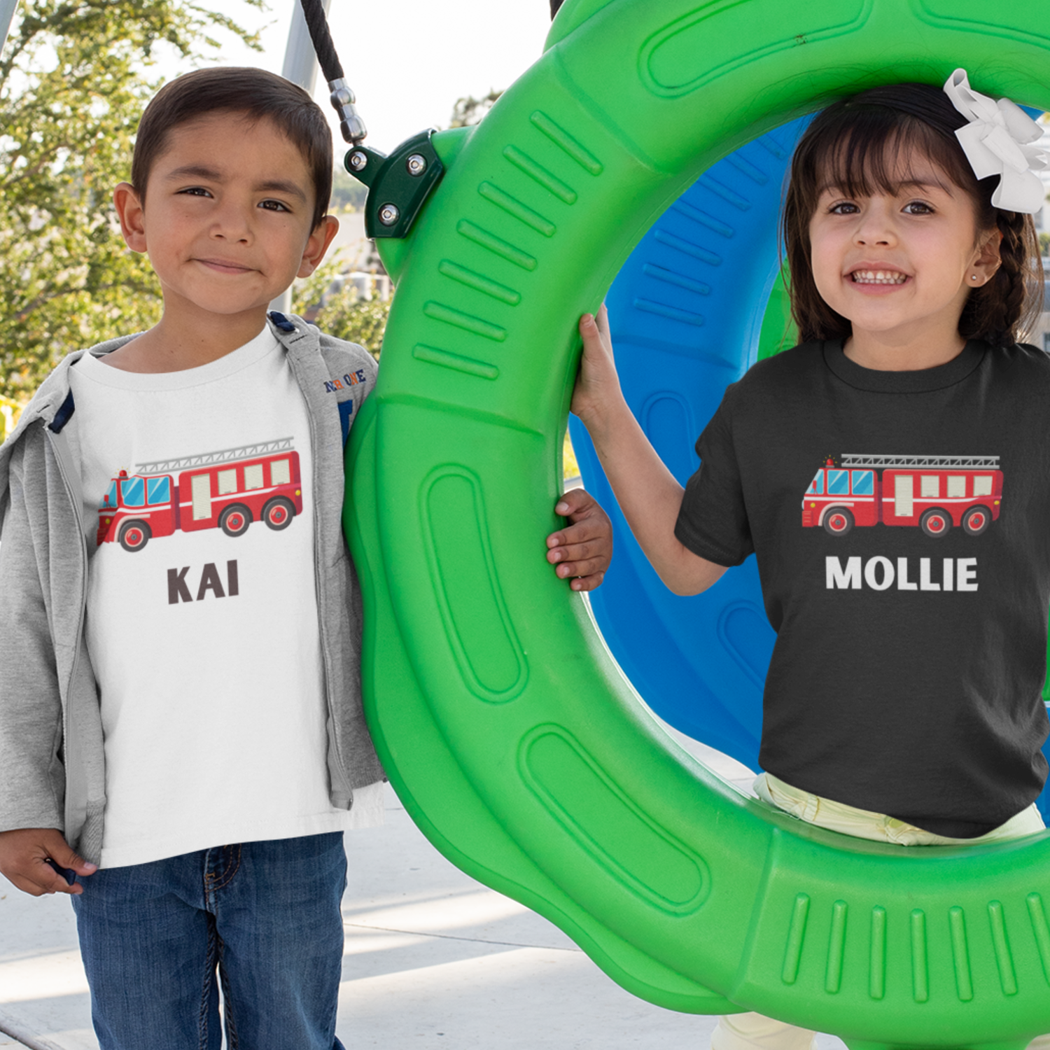 Boy and girl wearing personalised kids fire engine T-shirts with their names printed under a red fire truck graphic at a playground.