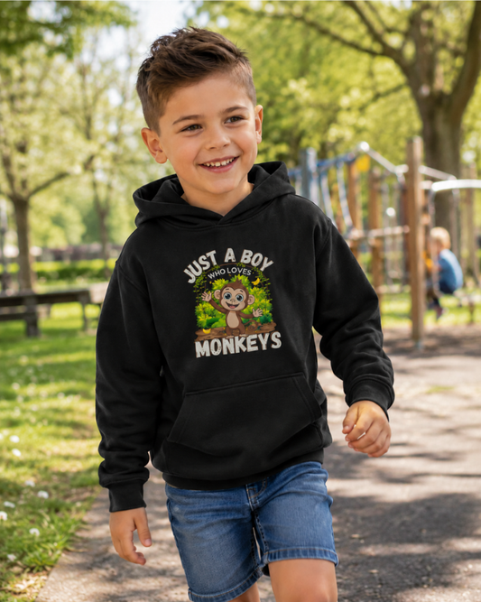 Happy young boy wearing a black “Just A Boy Who Loves Monkeys” kids hoodie and denim shorts walking through a sunny park playground, featuring a colourful monkey graphic print on the front.