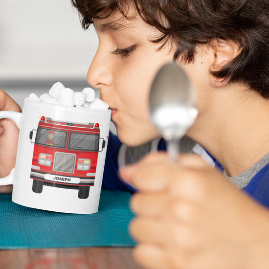Child drinking hot chocolate with marshmallows from a white mug featuring a red fire engine and personalised name on the number plate.