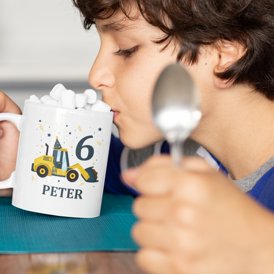 A child is sitting at a table, holding a spoon in his hand, while sipping from a white mug that has a printed birthday-themed design of a yellow digger wearing a party hat with the numbers "6" and "Peter" written underneath.