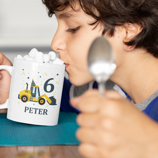 A child is sitting at a table, holding a spoon in his hand, while sipping from a white mug that has a printed birthday-themed design of a yellow digger wearing a party hat with the numbers 