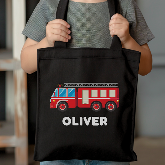 A boy holding a black tote bag with printed fire truck and his name printed underneath the truck