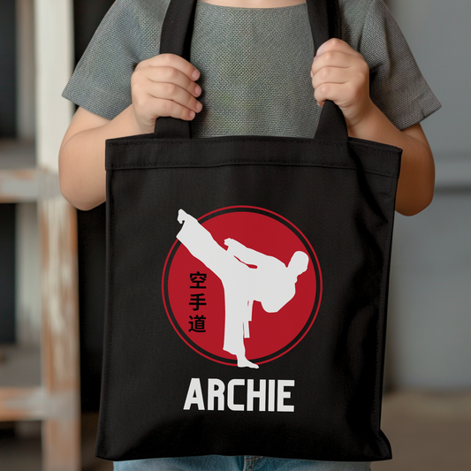 Child holding a black personalised karate tote bag with white high-kick silhouette, red circle emblem and name Archie printed underneath