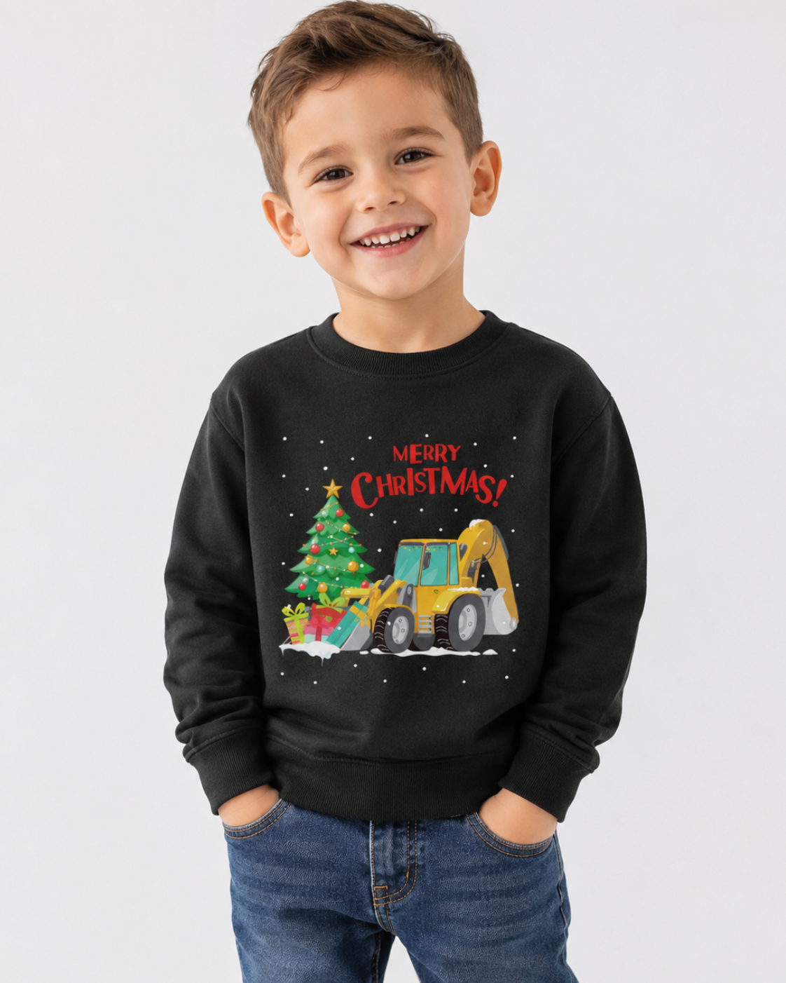 A young boy smiling and wearing a black crewneck sweatshirt with a colourful graphic of a Christmas tree, gifts, and a yellow excavator under the text 'MERRY CHRISTMAS!'. He is also wearing blue jeans, set against a plain light grey studio background.