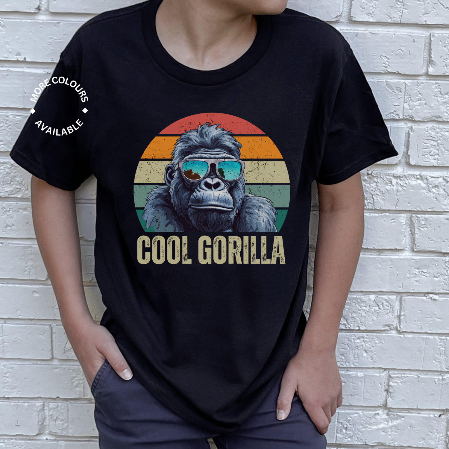 A young boy wearing a black short-sleeved t-shirt with a graphic of a cool gorilla in blue reflective sunglasses against a retro sunset background. Below the gorilla, the text 'COOL GORILLA' is printed. The boy stands in front of a white brick wall with his hands in his pockets."