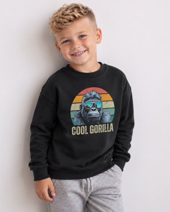 A young boy smiling while wearing a black crewneck sweatshirt with a retro-style graphic of a gorilla wearing sunglasses and the text 'COOL GORILLA' printed below it, paired with light grey sweatpants against a plain light grey wall.