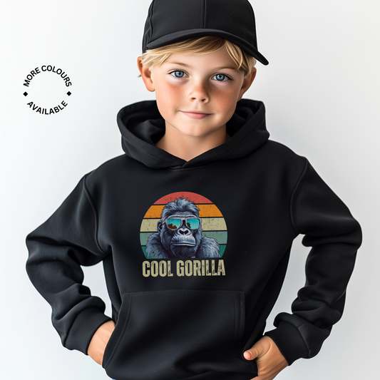 A young boy with blonde hair and a black baseball cap wears a black pullover hoodie featuring a circular graphic of a cool gorilla in blue reflective sunglasses against a retro sunset background, with 'COOL GORILLA' text below. 