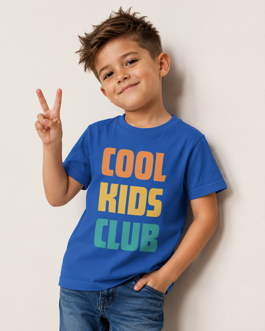 A young boy with styled brown hair wearing a blue t-shirt with 'COOL KIDS CLUB' printed in orange, yellow, and green text, posing against a light beige wall in studio setting.