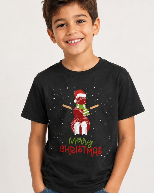 A smiling young boy wearing a black short-sleeve t-shirt with a festive Christmas graphic of a snowman made from cricket balls, a red Santa hat, with 'Merry Christmas' written below, paired with blue jeans against a light grey background.