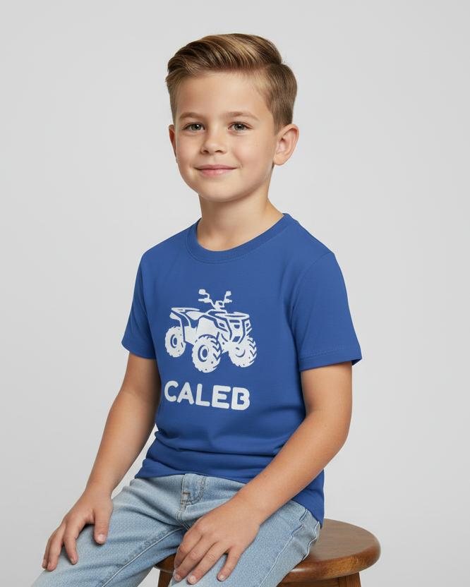 Boy sitting on a stool wearing a royal blue personalised kids t-shirt with a white quad bike graphic and the name “Caleb”, studio background.