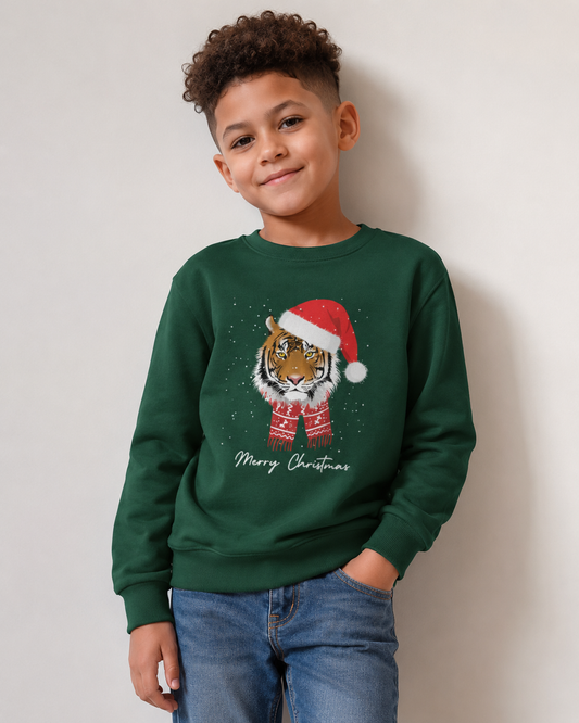 A young boy wearing a dark green crewneck sweatshirt with a festive graphic of a tiger in a red Santa hat and scarf, with 'Merry Christmas' written below, paired with blue jeans against a plain light gray wall.