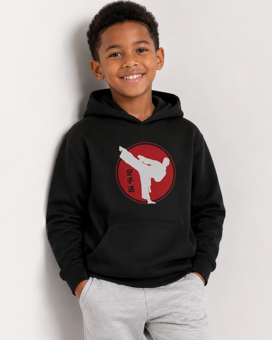 A smiling young boy wearing a black pullover hoodie with a red circular graphic and white martial arts silhouette on the chest, paired with light gray sweatpants, standing against a plain light gray wall.
